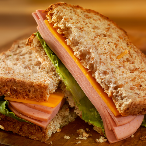 A close-up of a toasted whole wheat sandwich cut in half, filled with sliced bologna, cheese, and lettuce on a wooden surface.
