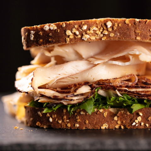 A close-up of a thick sandwich on dark bread with sliced turkey, and green lettuce, set against a dark background.