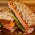 A close-up of a toasted whole wheat sandwich cut in half, filled with sliced bologna, cheese, and lettuce on a wooden surface.