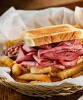 A close-up of a toasted sandwich filled with sliced corned beef and served with french fries in a woven basket lined with white paper, with a small side of ketchup.