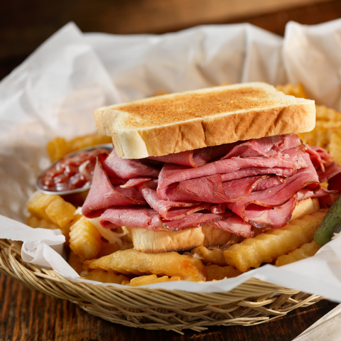 A close-up of a toasted sandwich filled with sliced corned beef and served with french fries in a woven basket lined with white paper, with a small side of ketchup.