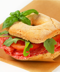 A bagel sandwich filled with sliced hard salami, tomato slices, and green leaves, placed on brown paper against a white background.