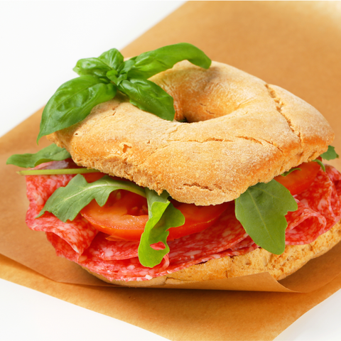 A bagel sandwich filled with sliced hard salami, tomato slices, and green leaves, placed on brown paper against a white background.