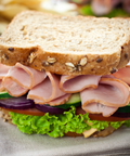 A close-up of a gourmet sandwich filled with sliced maple ham, cucumber, red onion, tomato, and lettuce on a seeded brown bread, set on a textured beige cloth with a blurred background.