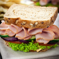 A close-up of a gourmet sandwich filled with sliced maple ham, cucumber, red onion, tomato, and lettuce on a seeded brown bread, set on a textured beige cloth with a blurred background.