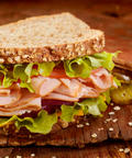 A close-up of a thick slice of whole wheat bread sandwiched with lettuce, sliced maple turkey, tomato, red onion, and pickles on a rustic wooden board.