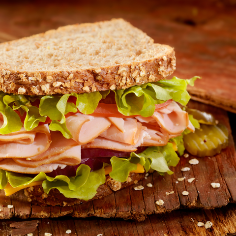 A close-up of a thick slice of whole wheat bread sandwiched with lettuce, sliced maple turkey, tomato, red onion, and pickles on a rustic wooden board.