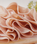 Thinly sliced chicken arranged in overlapping curls on a light wooden board with a small green herb garnish in the background.