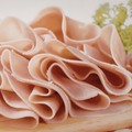 Thinly sliced chicken arranged in overlapping curls on a light wooden board with a small green herb garnish in the background.