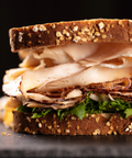 A close-up of a thick sandwich on dark bread with sliced turkey, and green lettuce, set against a dark background.