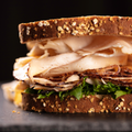 A close-up of a thick sandwich on dark bread with sliced turkey, and green lettuce, set against a dark background.