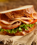 A close-up of a gourmet sandwich on a rustic wooden table, featuring a thick slice of white bread, sliced peppered turkey, tomato, and fresh green lettuce on a piece of burlap fabric.