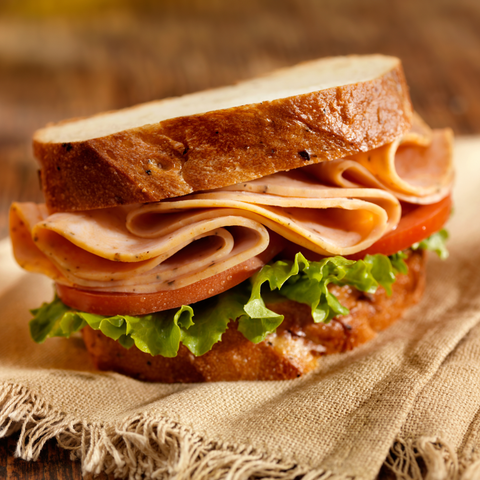 A close-up of a gourmet sandwich on a rustic wooden table, featuring a thick slice of white bread, sliced peppered turkey, tomato, and fresh green lettuce on a piece of burlap fabric.