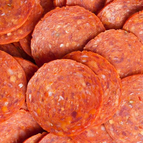A close-up pile of round pepperoni slices showing the textured, seasoned meat surface with visible spices and small flecks.