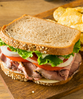 A large sandwich on a wooden board with potato chips nearby. The sandwich uses thick seeded bread, sliced roast beef, tomato, white onion, and green lettuce.
