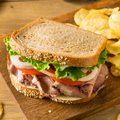 A large sandwich on a wooden board with potato chips nearby. The sandwich uses thick seeded bread, sliced roast beef, tomato, white onion, and green lettuce.