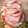 Slices of smoked ham layered over a sandwich with tomato and green lettuce on a piece of bread, set on a brown textured surface.